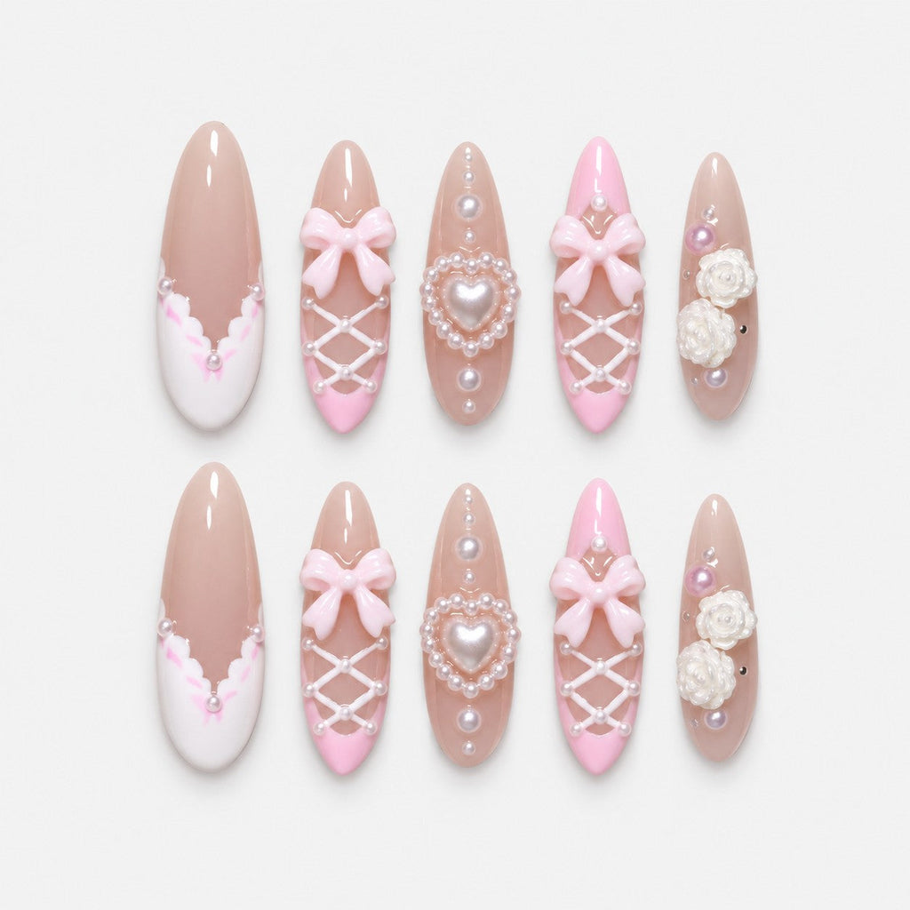 catalog: Long almond press-on nails in sheer nude with pink French tips, white corset lacing, pearls, sculpted pink bows and a pearl heart charm.