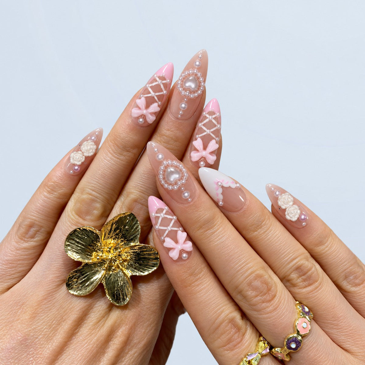 hand: Long almond press-on nails in sheer nude with pink French tips, white corset lacing, pearls, sculpted pink bows and a pearl heart charm.