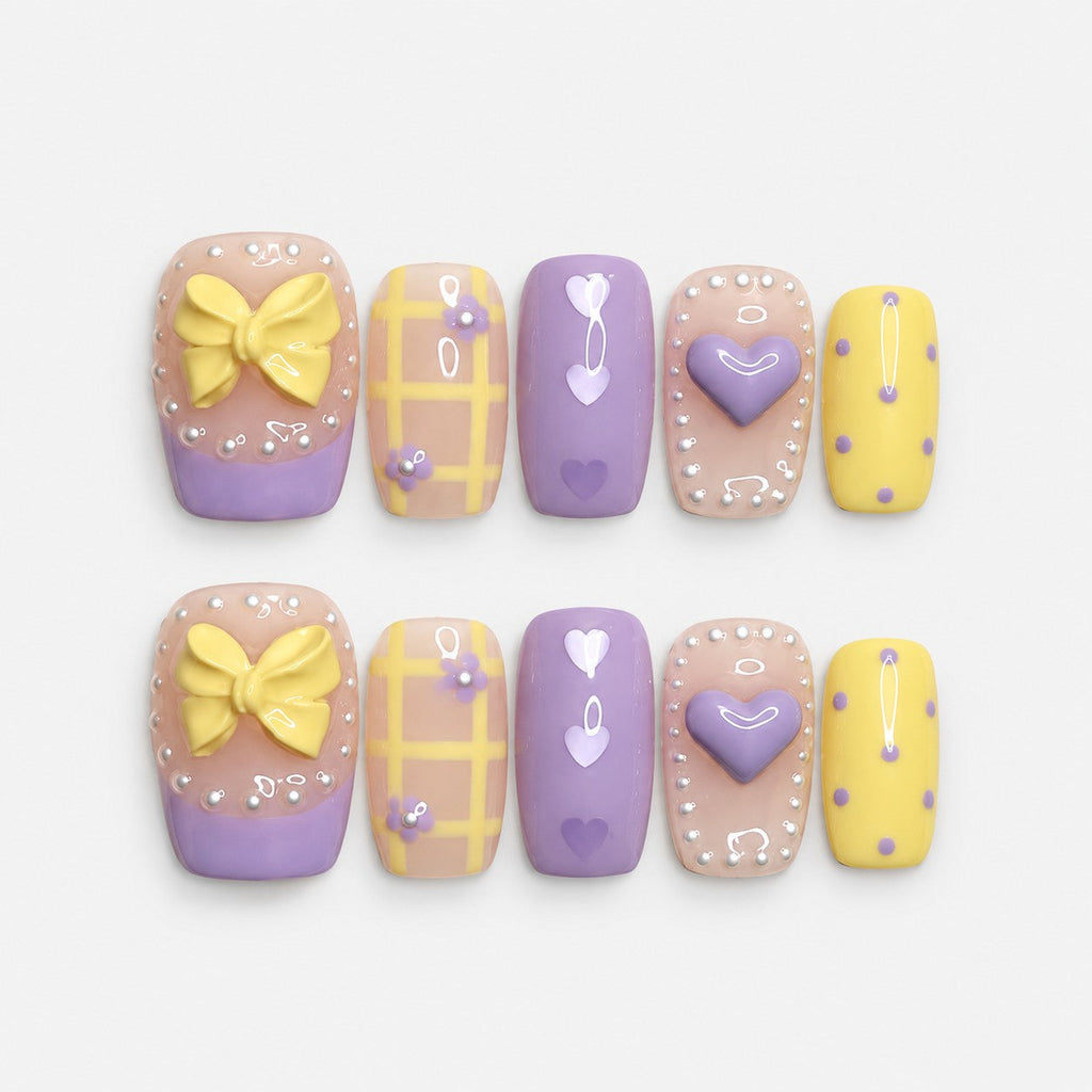 catalog: Short square press-on nails in lilac and pastel yellow with a sculpted bow, a raised heart, painted hearts, grid lines and white dot borders.