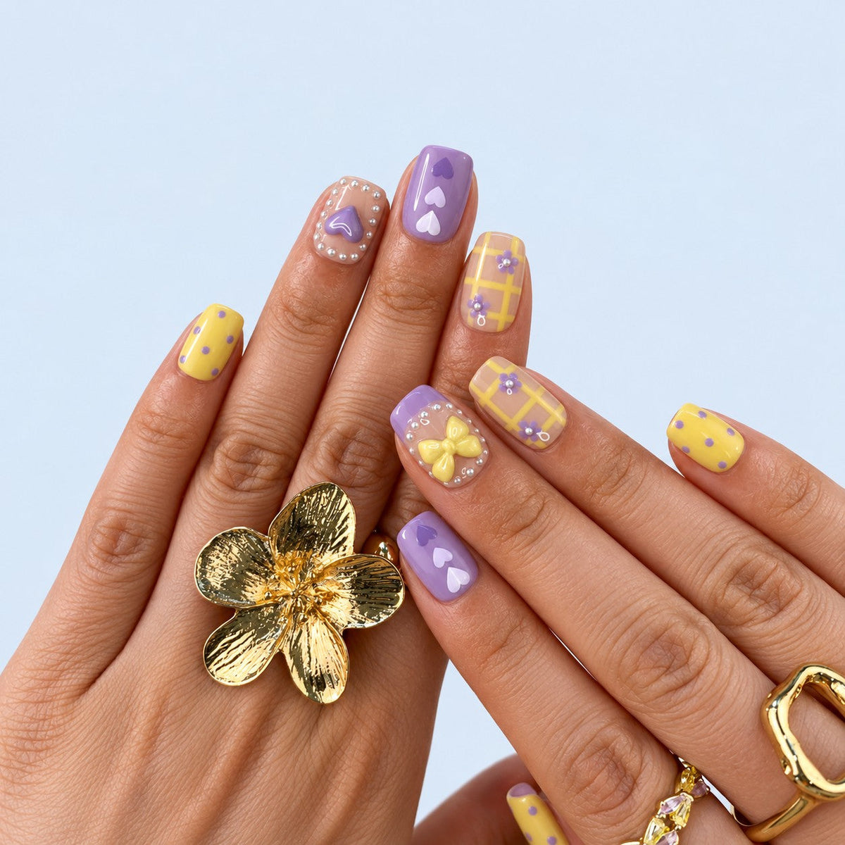 hand: Short square press-on nails in lilac and pastel yellow with a sculpted bow, a raised heart, painted hearts, grid lines and white dot borders.