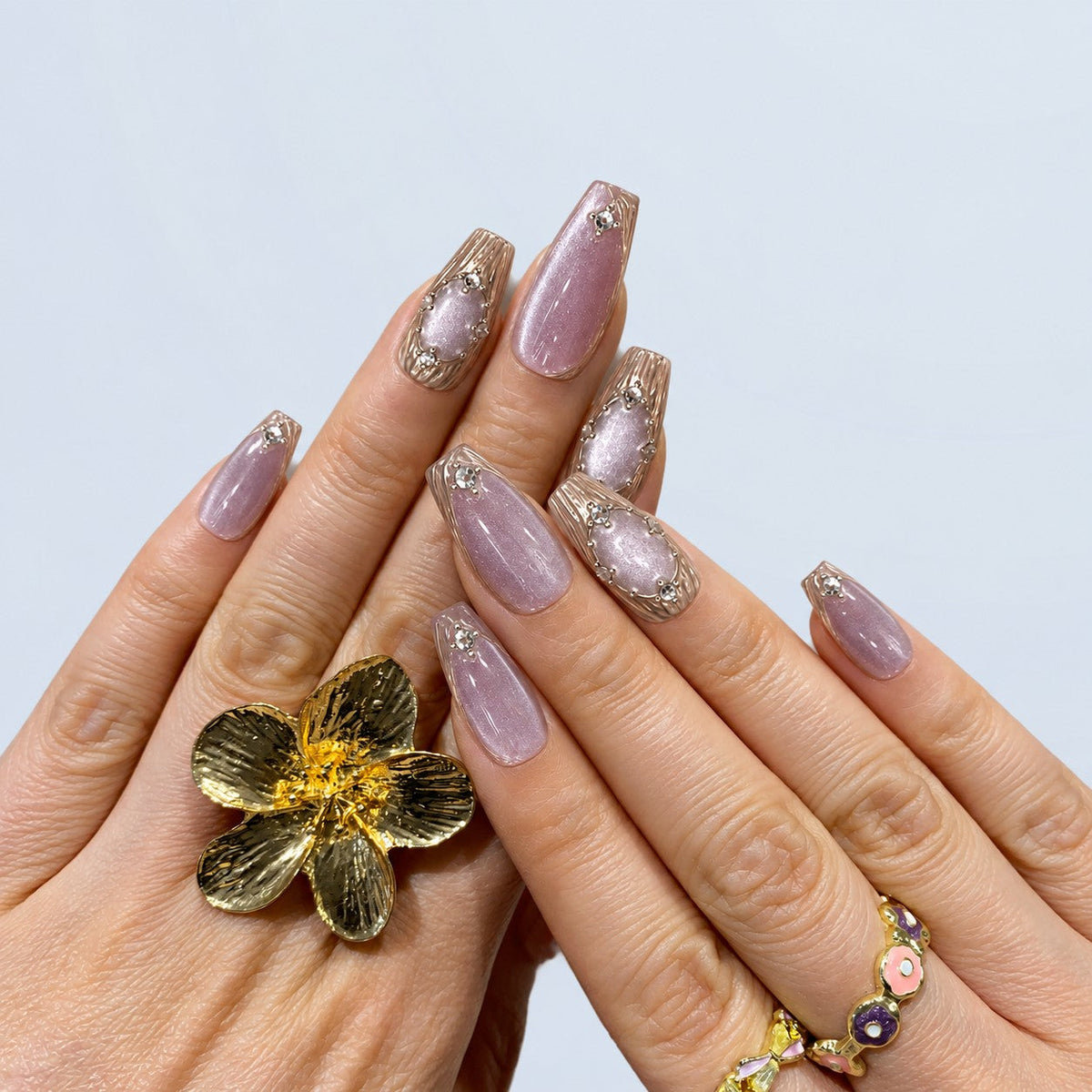 hand: Medium almond press-on nails in shimmering lilac cat-eye with sculpted chrome petal accents and small crystal clusters.