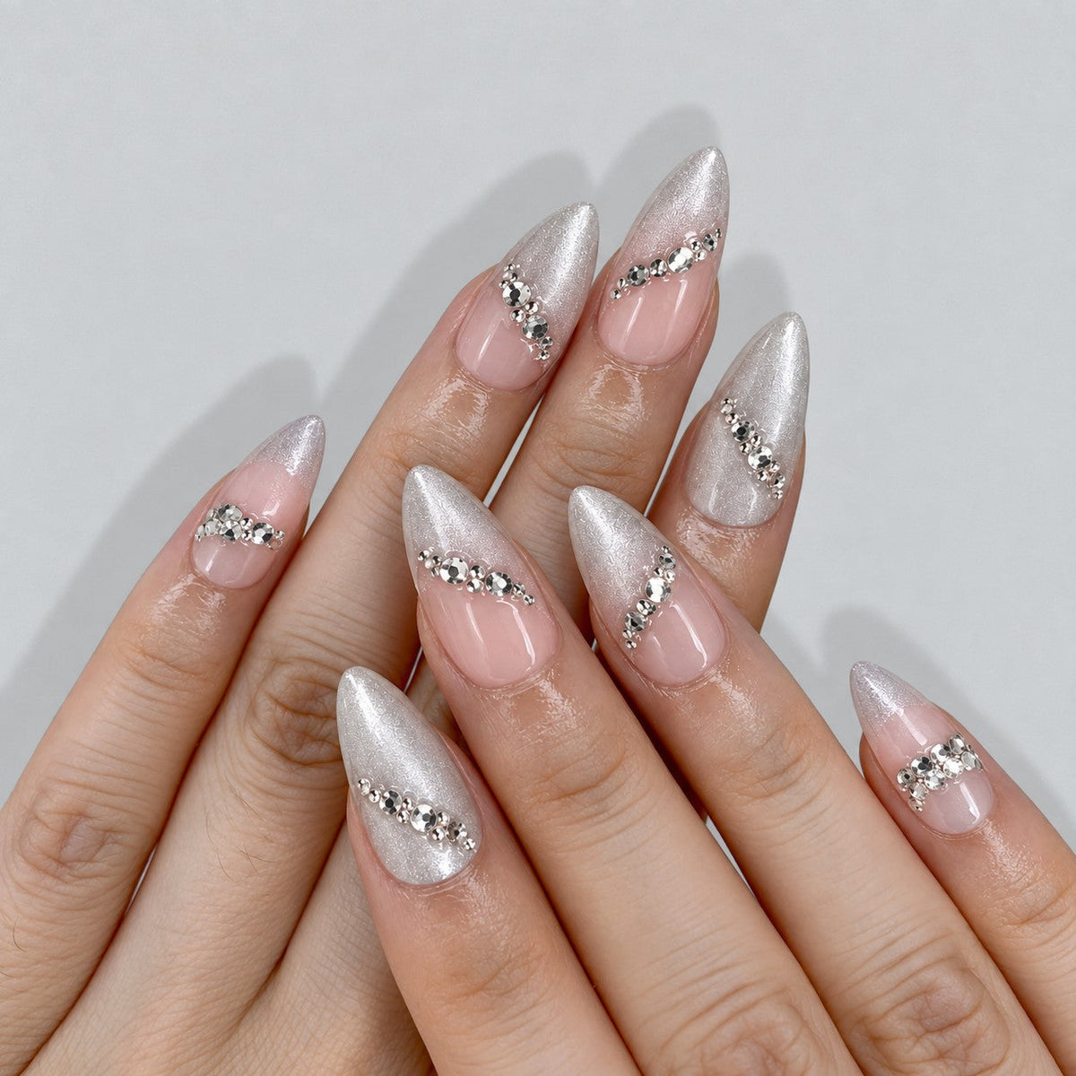 hand: Medium almond press-on nails in nude with pearly silver cat-eye shimmer, crystal accents along the edges and two large faceted stones.