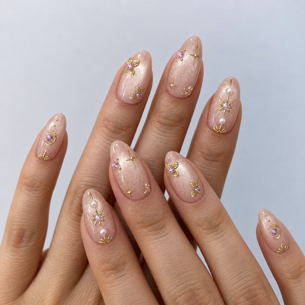 hand: Medium almond press-on nails in shimmering pearly nude with gold star linework, pearls and a lilac gemstone flower.
