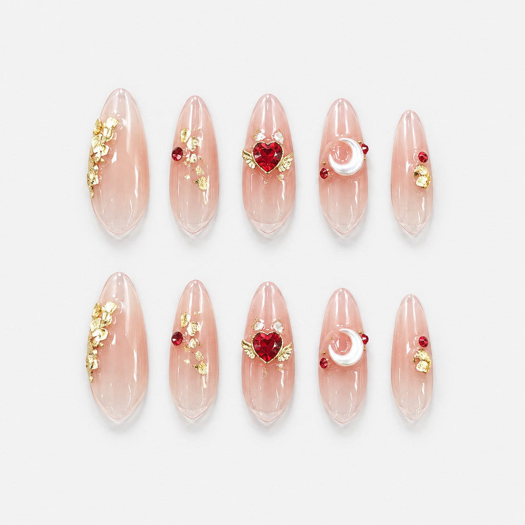 catalog: Long almond press-on nails in clear nude with gold leaf flecks, a red heart crystal with gold wings, and a pearl crescent moon charm.