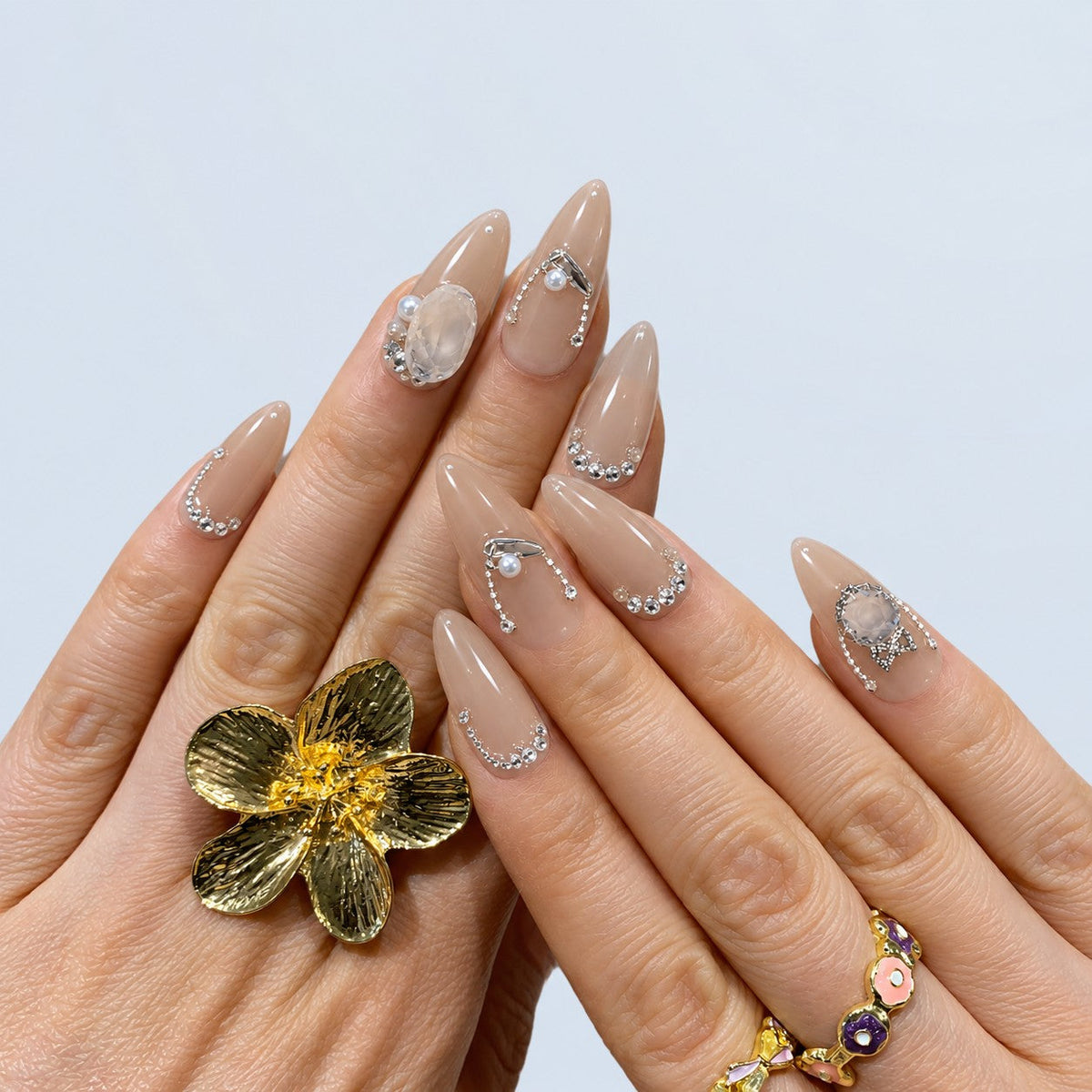 hand: Long almond press-on nails in glossy greige nude with silver rhinestone curves, a bow charm, a large clear crystal and small pearls.