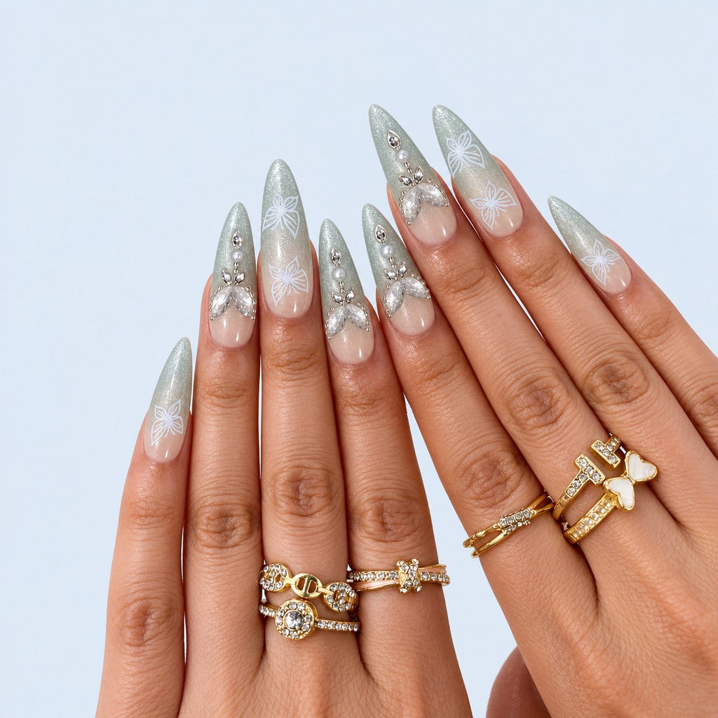 hand: Long almond press-on nails with nude to sage green cat-eye gradient, white painted butterflies, crystals, pearls and a raised gold and stone butterfly charm.