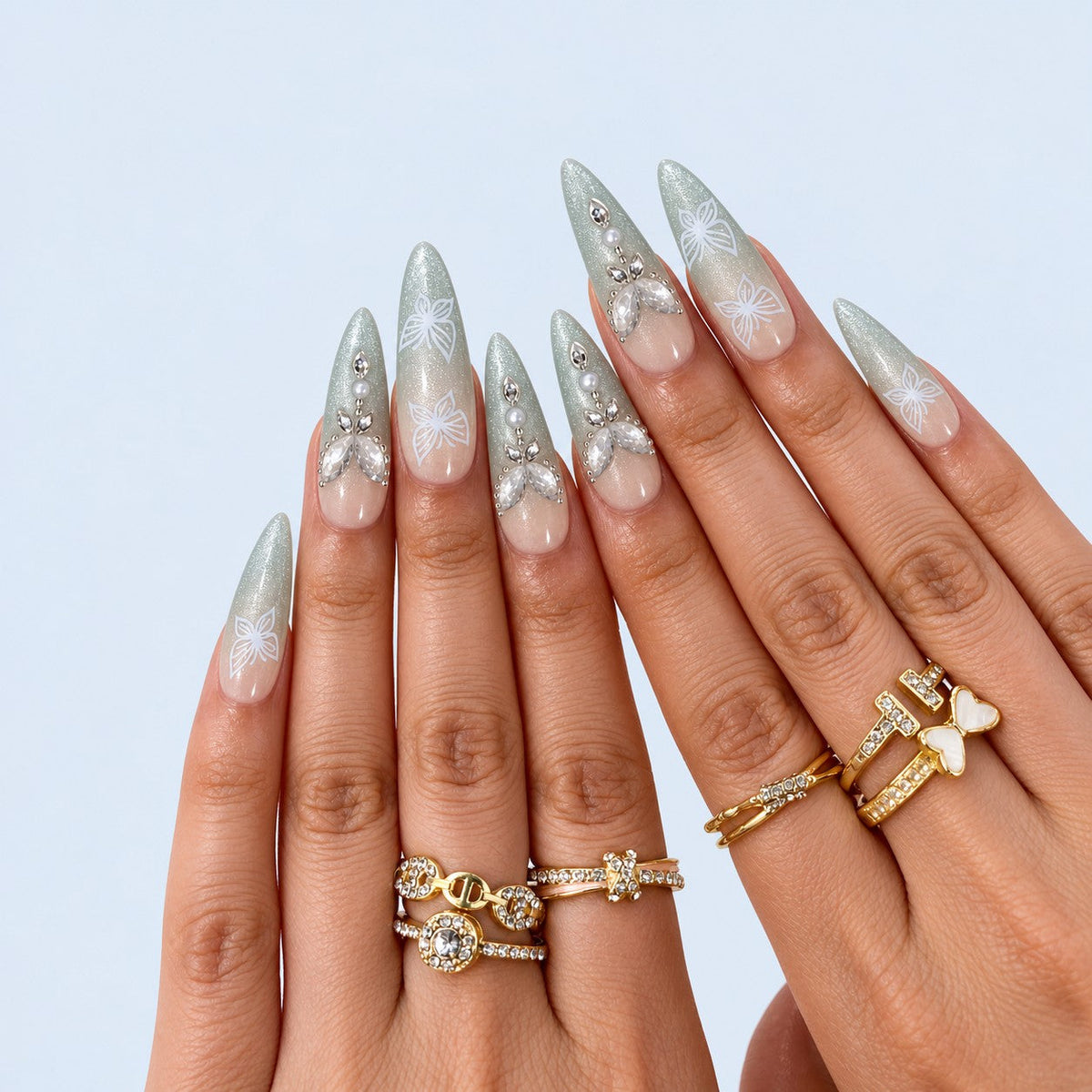 hand: Long almond press-on nails with nude to sage green cat-eye gradient, white painted butterflies, crystals, pearls and a raised gold and stone butterfly charm.