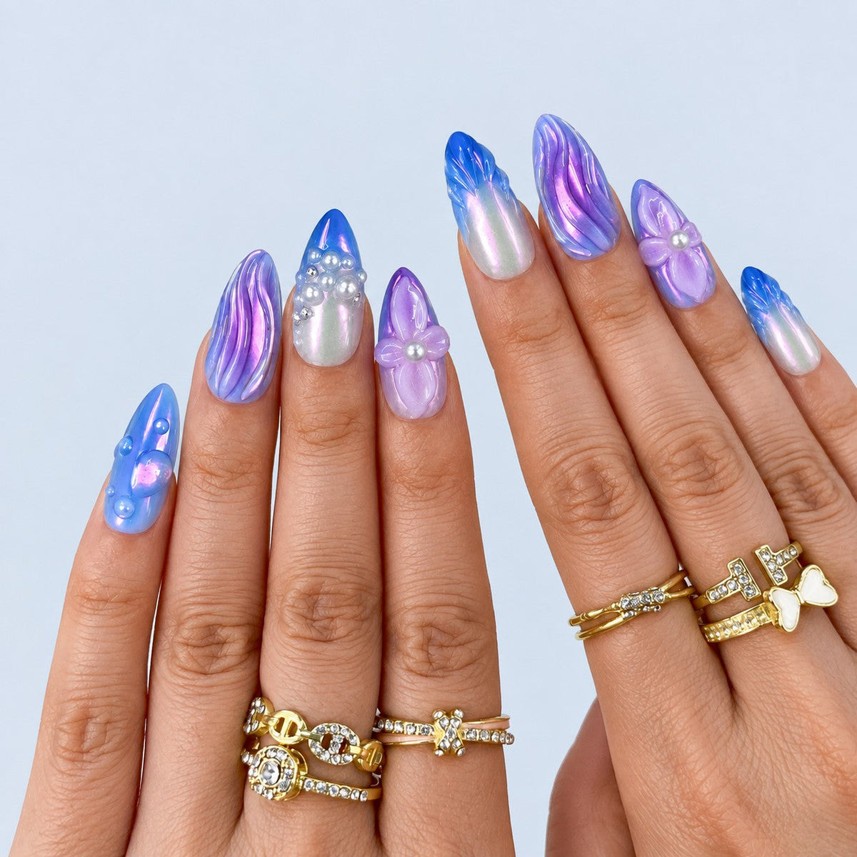 hand: Long almond press-on nails in lilac, violet and blue chrome with sculpted ripples, raised flowers with pearl centres and clustered pearl accents.