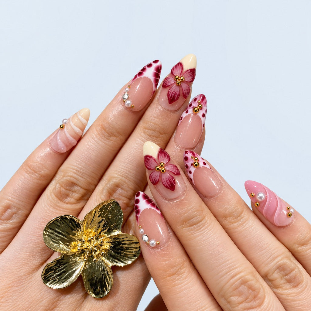 hand: Medium almond press-on nails in soft pink with raised burgundy 3D flowers, wine-coloured speckled tips, carved wave texture, pearls and gold beads.