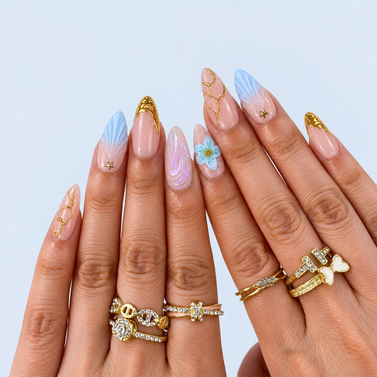 hand: Almond press-on nails in soft pink with sculpted blue wings, a pale blue flower, gold vein lines, gold star studs and gold-dipped tips.