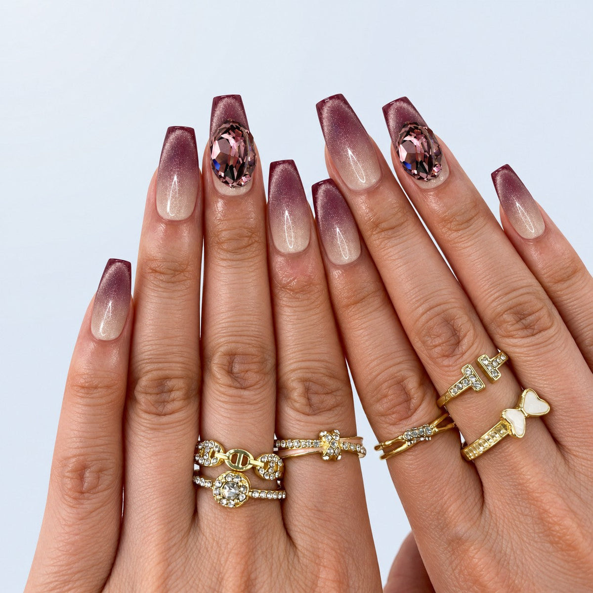 hand: Medium coffin-shaped press-on nails with a nude to plum cat-eye gradient and a large pink faceted crystal on one nail.
