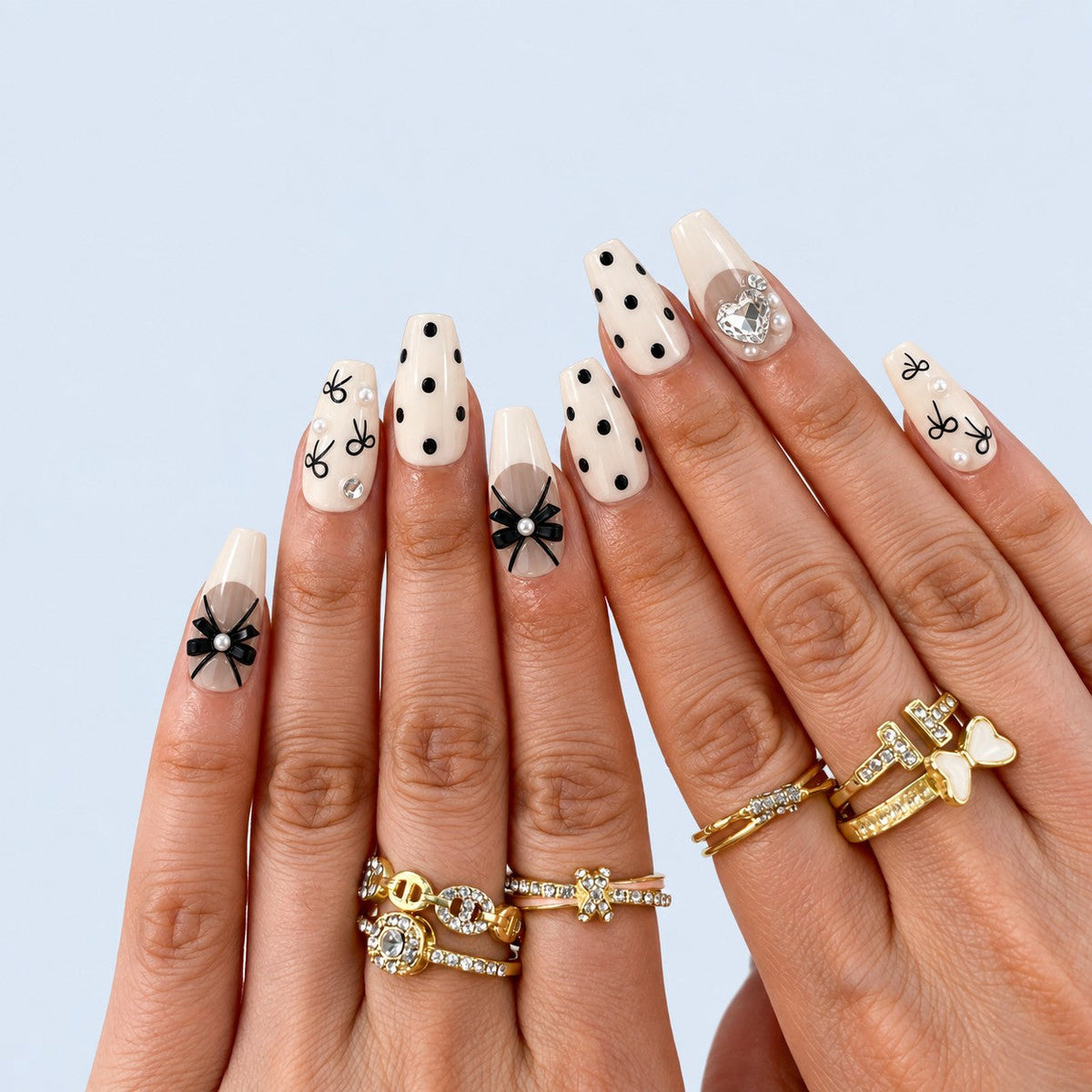 hand: Medium coffin press-on nails in cream with sheer French tips, black satin bows, black polka dots and clear heart-shaped crystals.