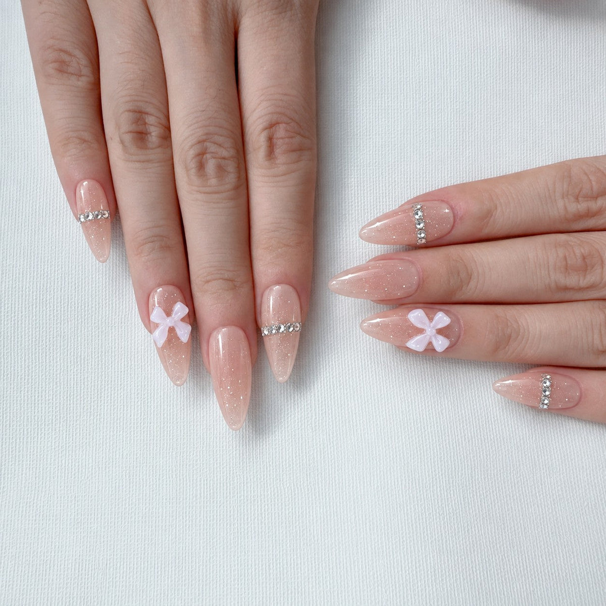hand: Long almond press-on nails in sheer pink with a row of small crystals along the cuticle line and a white 3D bow on the ring finger.