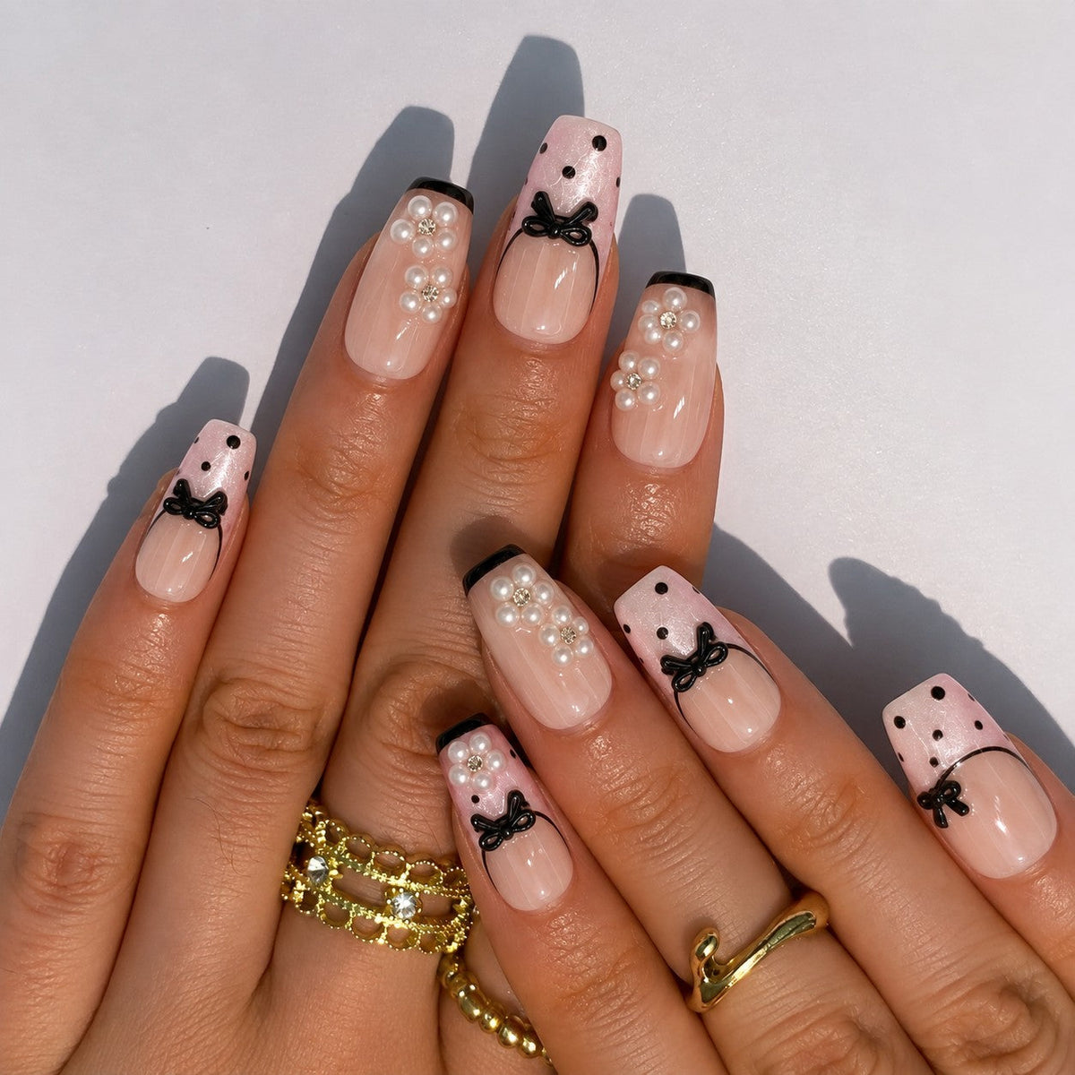 hand: Medium square press-on nails in sheer blush pink with thin black French tips, black dots, raised black bows and pearl flower clusters.