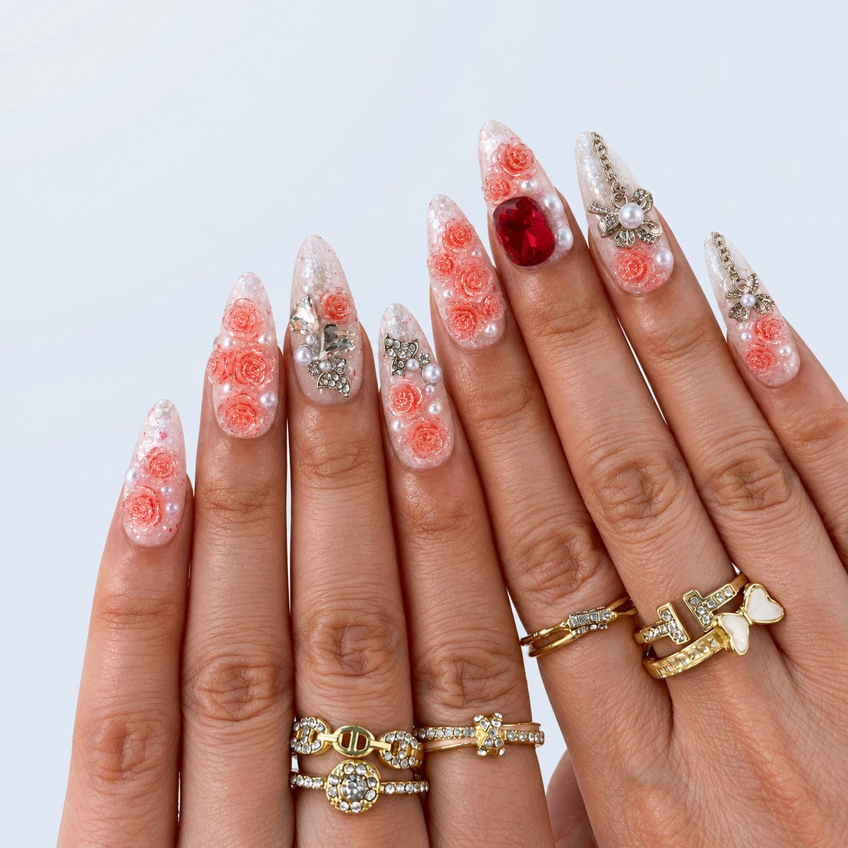 hand: Long almond press-on nails with clear bases, sculpted coral rose flowers, pearls, clear and red crystals, and silver bow charms with chains.