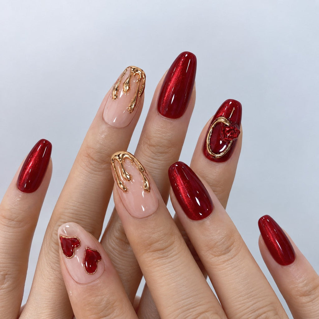 hand: Medium almond press-on nails in deep red cat-eye with a sculpted rose in a gold ring, a gold-marbled nude nail and two glossy red hearts.