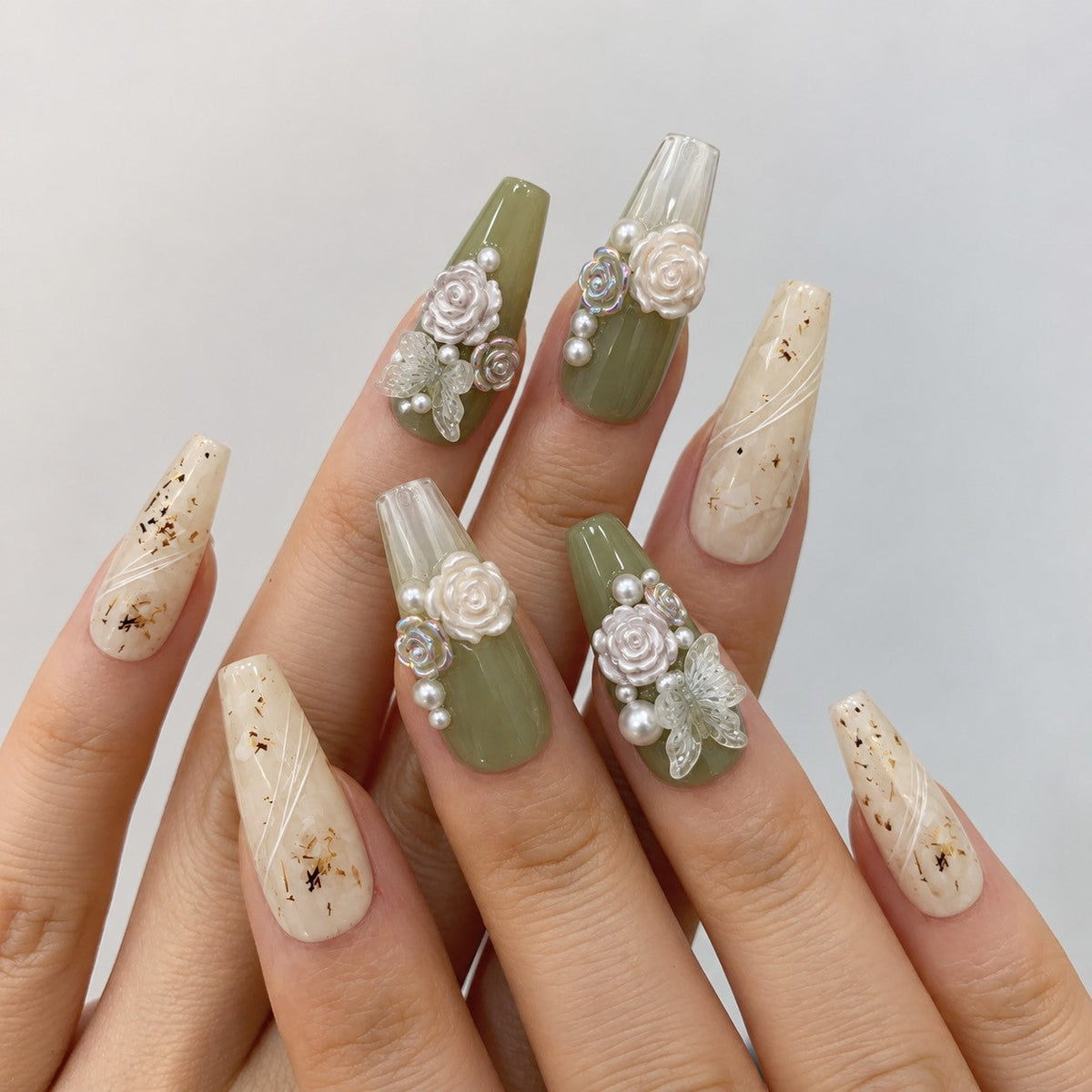 hand: Long coffin press-on nails in sage green and milky white with hand-sculpted white roses, pearls and sheer butterflies, plus clear nails holding dried flower flecks.
