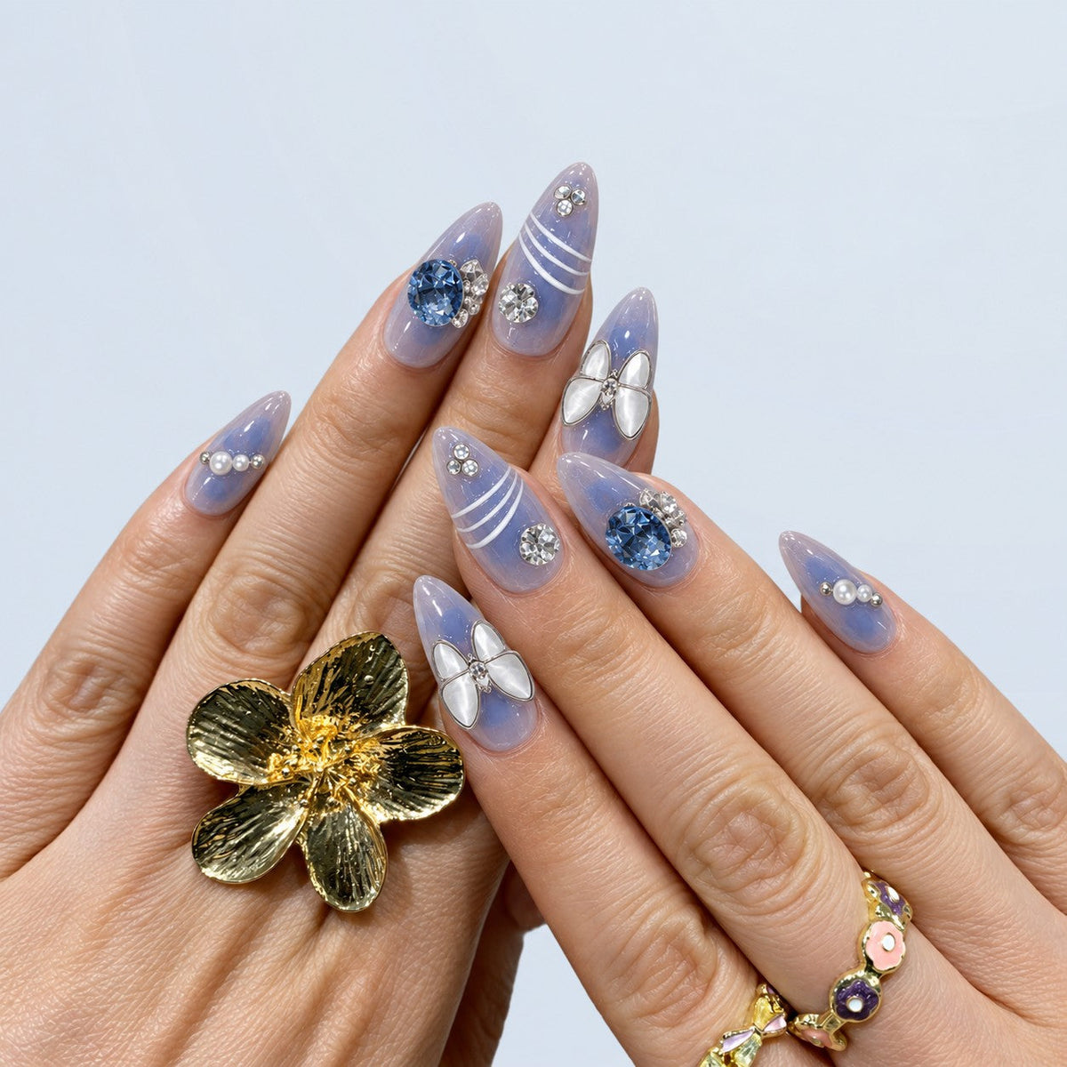 hand: Long almond press-on nails in translucent blue and lilac marble with a white butterfly charm, a large blue gem, white curved lines and pearl details.