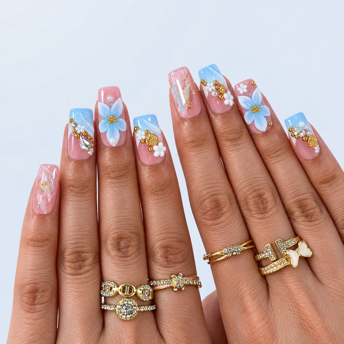 hand: Medium square press-on nails in pink to blue gradient with sculpted white and blue flowers, gold shell charms, pearls and gold leaf marbling.