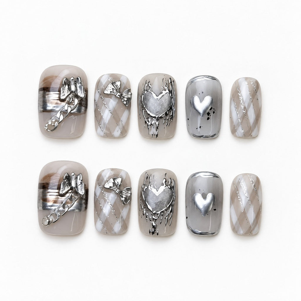 catalog: Short square press-on nails in smoky grey with silver cat-eye shimmer, chrome hearts, a sculpted bow and a tiny chain.
