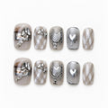 catalog: Short square press-on nails in smoky grey with silver cat-eye shimmer, chrome hearts, a sculpted bow and a tiny chain.