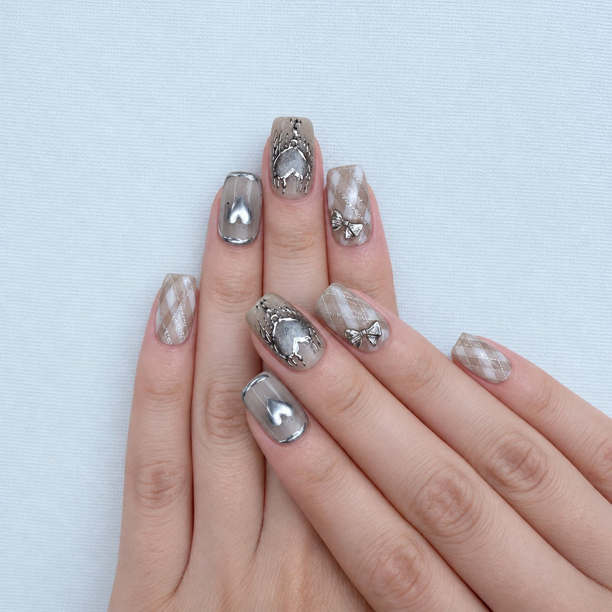 hand: Short square press-on nails in smoky grey with silver cat-eye shimmer, chrome hearts, a sculpted bow and a tiny chain.