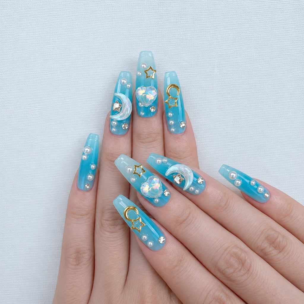 hand: Long coffin press-on nails in ocean blue fading to white, with sculpted mermaid tails, gold moon and star charms, crystals and pearls.