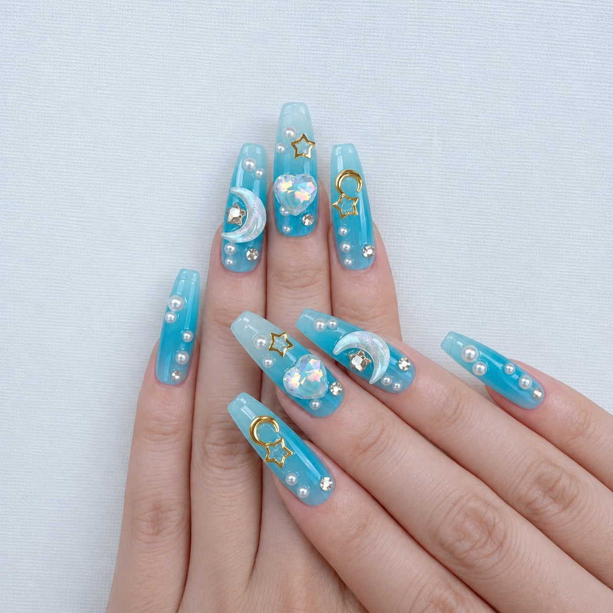 hand: Long coffin press-on nails in ocean blue fading to white, with sculpted mermaid tails, gold moon and star charms, crystals and pearls.