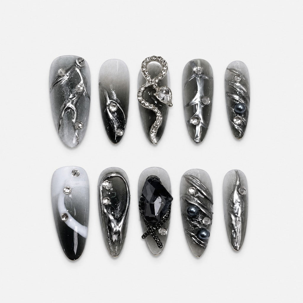 catalog: Long almond press-on nails in a smoky grey to black gradient with raised liquid chrome swirls, crystals, grey pearls and a rhinestone snake charm.