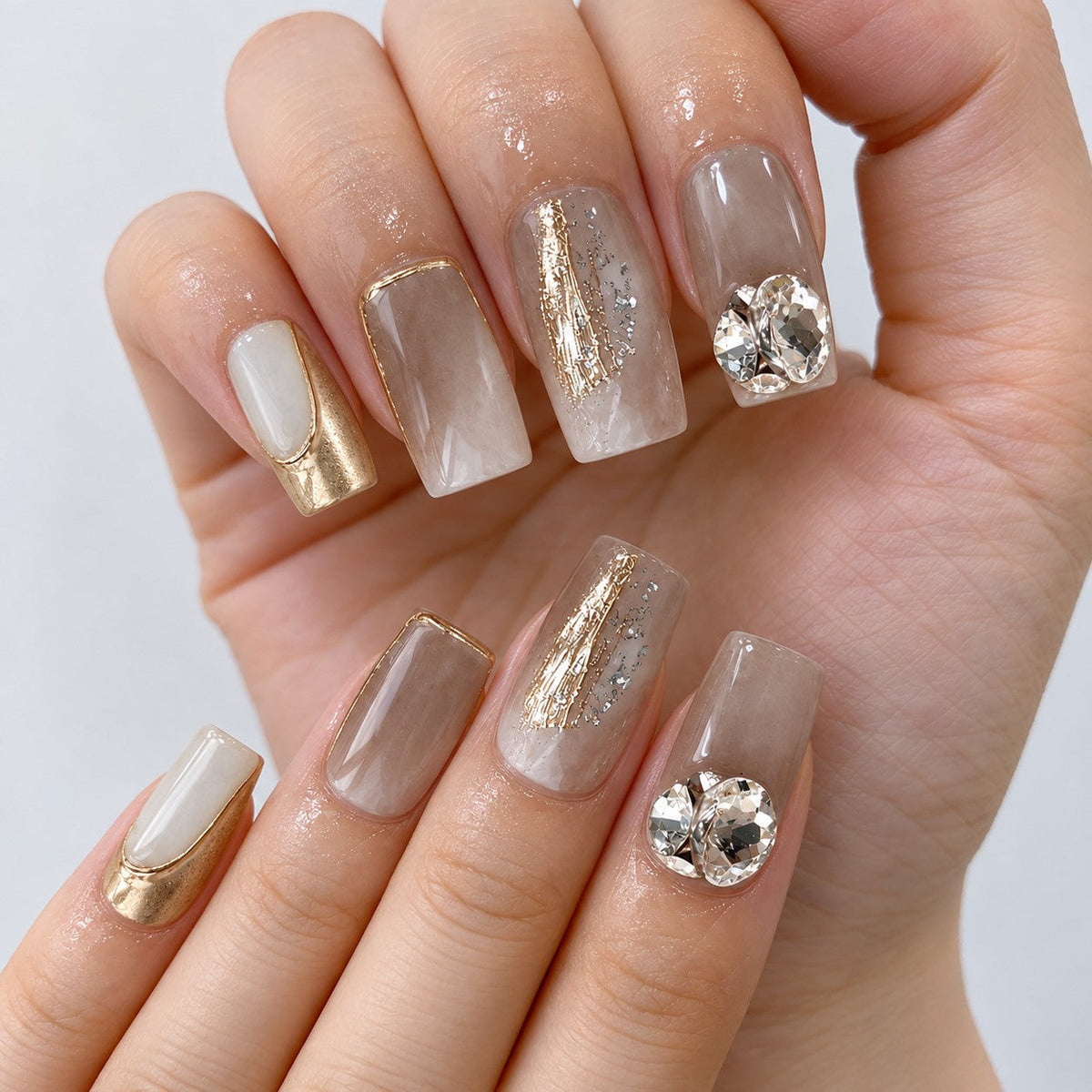 hand: Medium square press-on nails in smoky taupe marble with gold outlines, a mirrored gold nail, silver foil detail and large clear and champagne crystals.