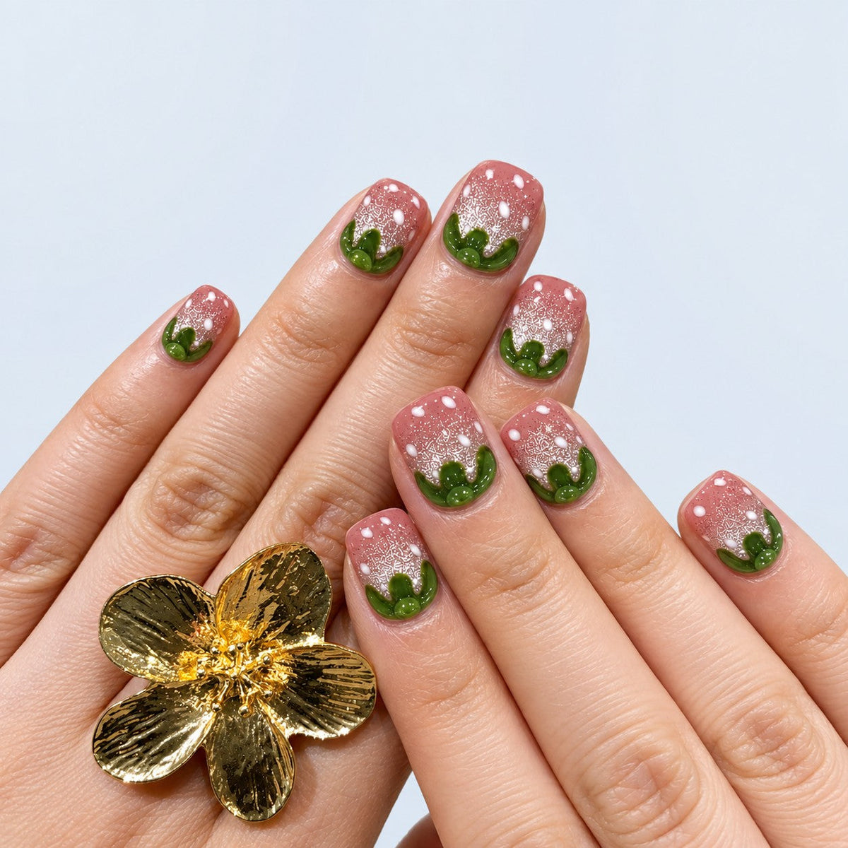 hand: Short square press-on nails shaped like strawberries, pink shimmer bases with raised white seed dots and sculpted glossy green tops.