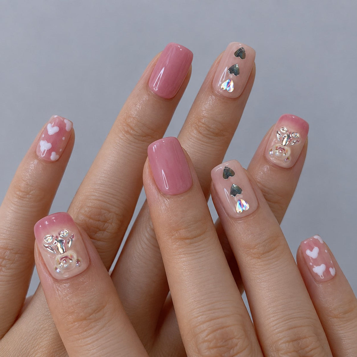 hand: Short square press-on nails in a pink to nude gradient with sculpted crystal bear charms, a silver heart and small white hearts.