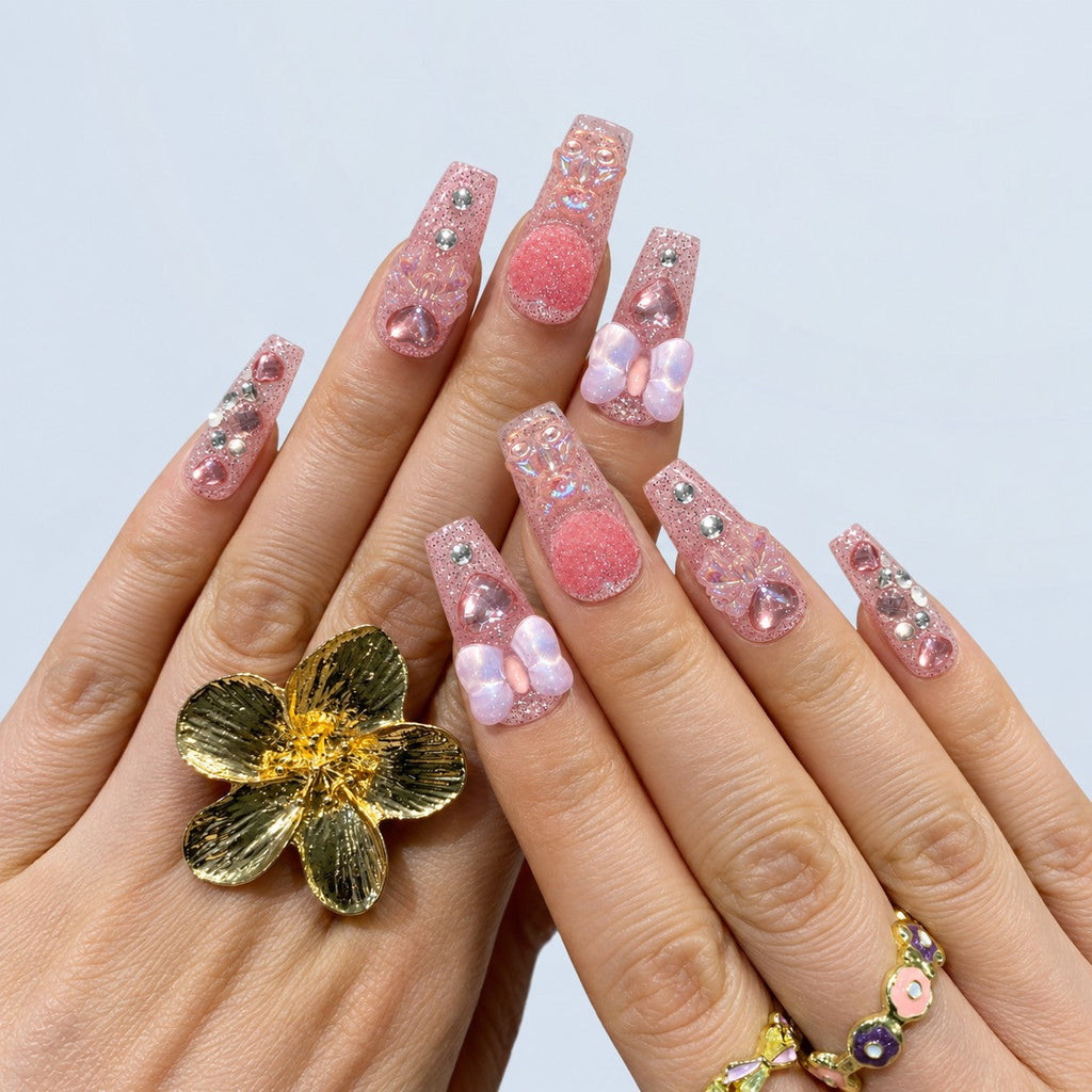 hand: Long square press-on nails in sheer pink glitter with sculpted teddy bear charms, iridescent bows, glass hearts and small rhinestones.