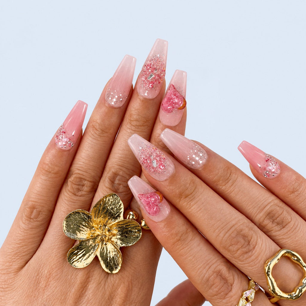 hand: Long coffin press-on nails in sheer pink with a raised glitter heart, silver beading and crystal grit at the cuticle.
