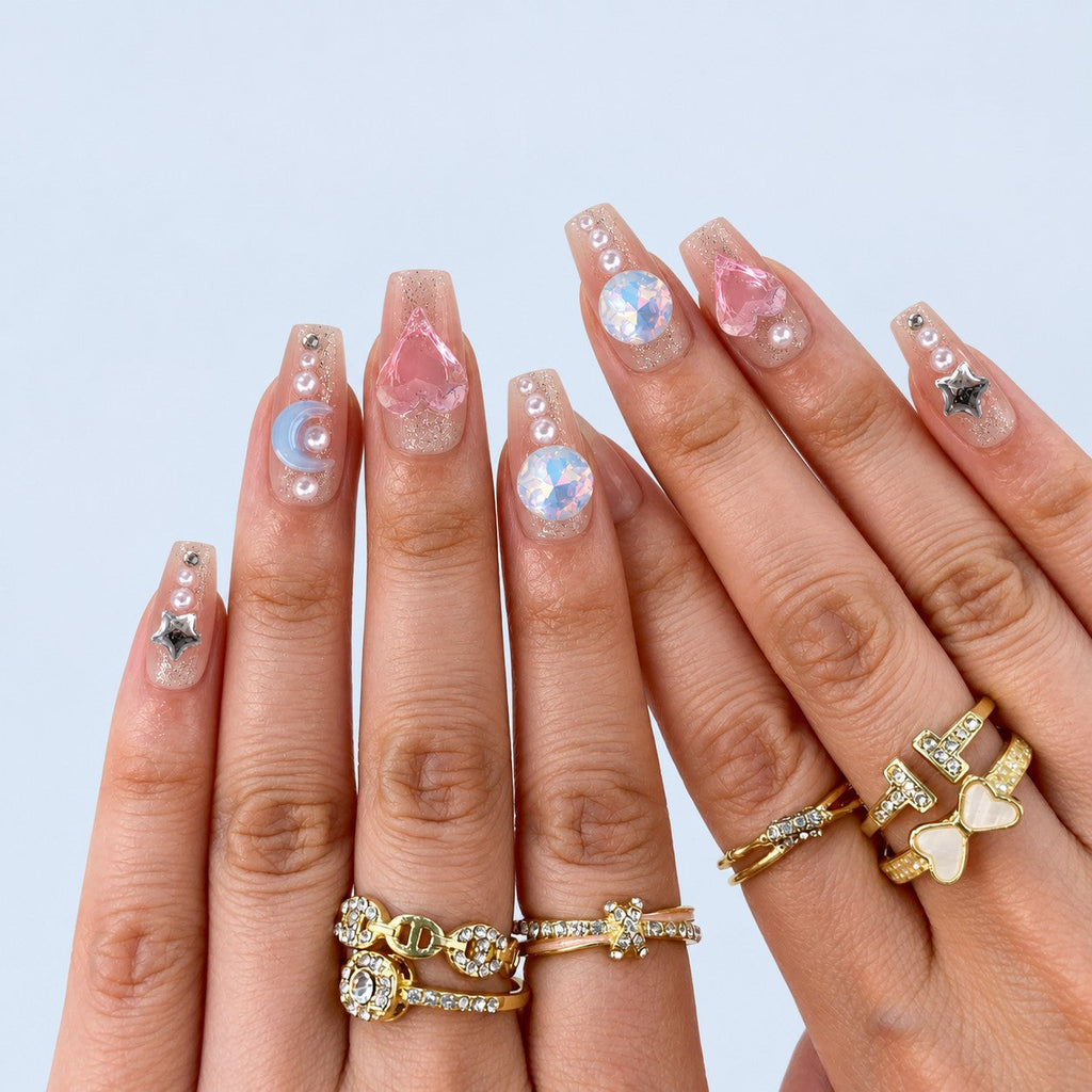 hand: Medium coffin press-on nails in sheer pink with cuticle glitter and charms including a heart, opal stone, butterfly, blue crescent, chrome stars and pearls.