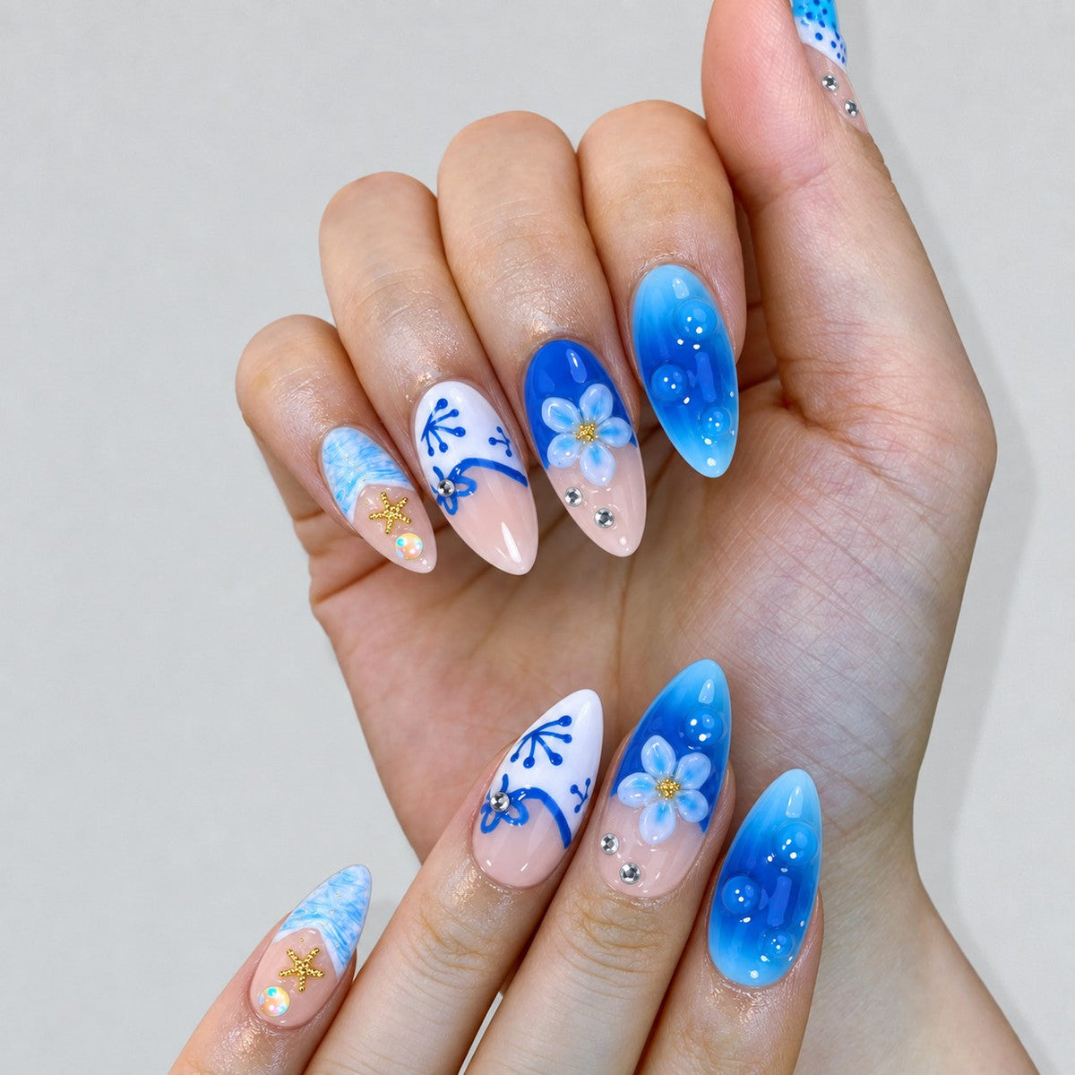 hand: Medium almond press-on nails in cobalt and aqua over sheer pink with hand-painted white coral, sculpted flowers, gold starfish and crystals.