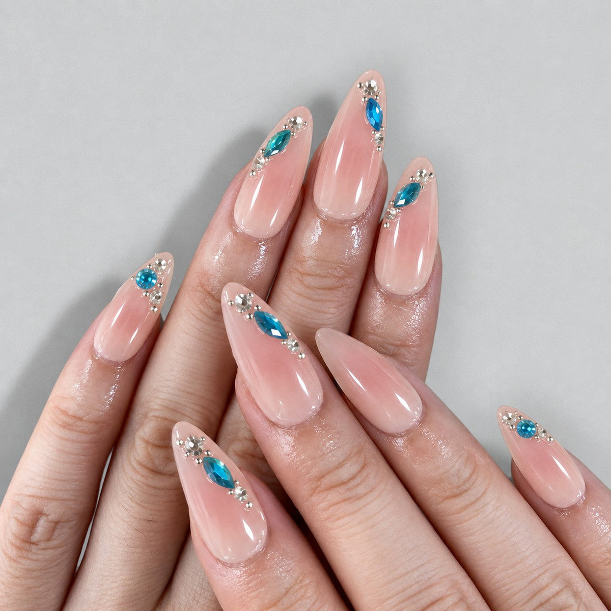 hand: Long almond nails in milky pink with glossy finish and blue teardrop gems set in silver along the tips.