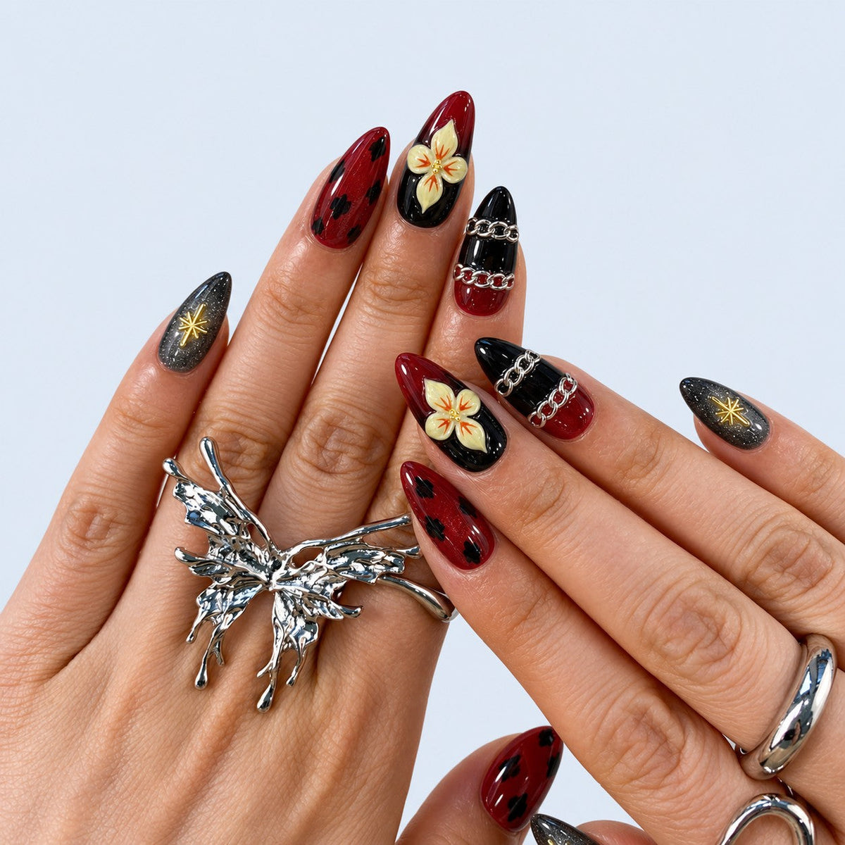 hand: Medium almond press-on nails in dark wine red and black with painted cream orchids, black spots, a gold star charm and raised silver chains.