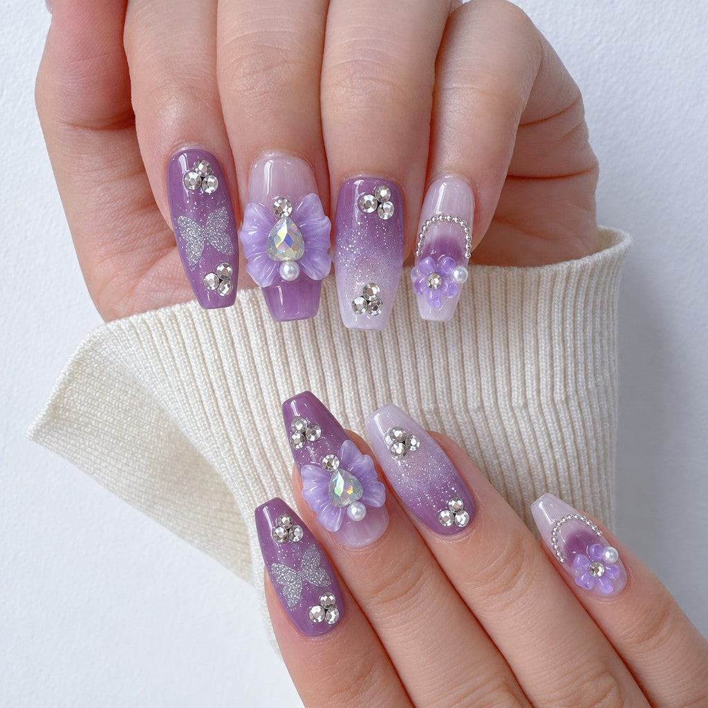 hand: Medium coffin press-on nails in white to lilac ombre with sculpted purple flowers, silver glitter and clear rhinestones.